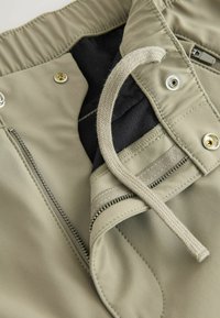 Gaiši bēšas bikses ar rāvējslēdzēju, auklu un sprādzīti aizdari, mīkstu melnu oderi un kontrastējošām šuvju detaļām.