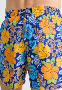 Blå svøm shorts med livlige oransje og turkise blomster mønstre, med elastisk livlinning og en merkeetikett bak.