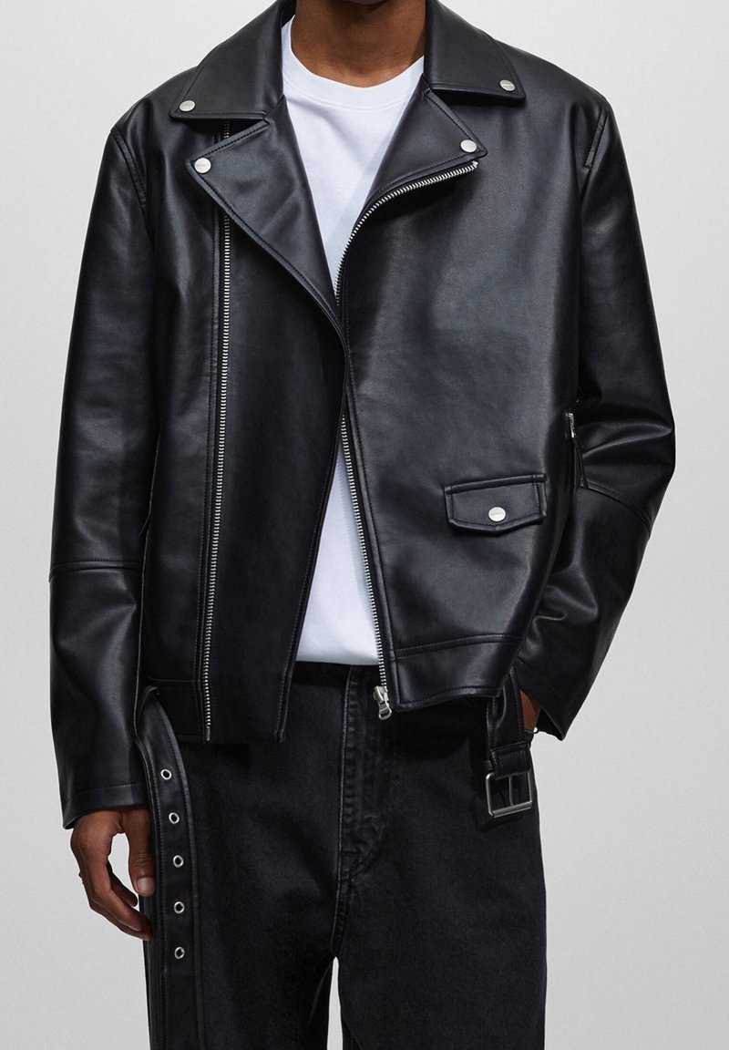 Chaqueta de cuero negra con diseño de motorista, cremalleras plateadas, botones de presión y un bolsillo lateral; llevada sobre una camiseta blanca y jeans oscuros.