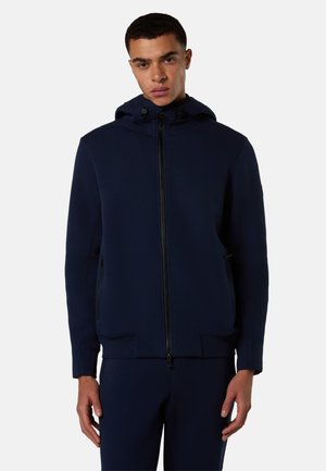 Giovane uomo che indossa una giacca con cappuccio e zip blu scuro e pantaloni abbinati, in piedi davanti a uno sfondo chiaro e semplice.