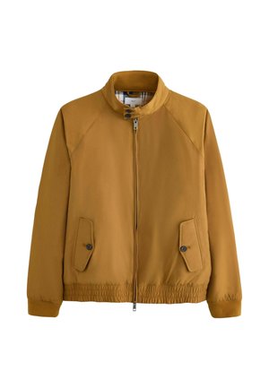 Mann trägt eine senfgelbe Jacke mit Reißverschluss, graue strukturierte Hose und braune Leder-Schnürschuhe, steht vor einem beigen Hintergrund.