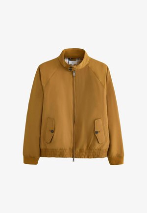 Mann trägt eine senfgelbe Jacke mit Reißverschluss, graue strukturierte Hose und braune Leder-Schnürschuhe, steht vor einem beigen Hintergrund.
