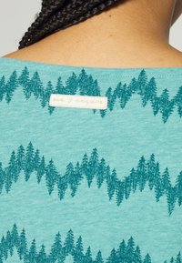 Turkusowa koszula z krótkim rękawem z teksturowanym wzorem zielonego drzewa. W tylnej części dekoltu znajduje się naszywka z napisem "kochamy wegan".