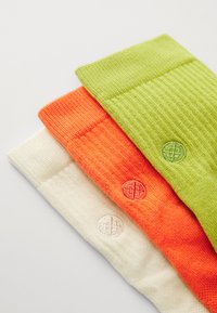 Three pairs of knitted socks: green, orange, and white, each with ribbed texture and circular embroidered logos near the cuffs.