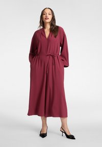 Bordowa sukienka kopertowa z długimi rękawami, głębokim dekoltem w serek i zawiązanym w pasie paskiem. Wykonana z gładkiego materiału, charakteryzuje się luźną, zwiewną sylwetką.