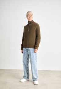 Muž v béžové pletené čepici, hnědém svetr s zipem a žebrovaným vzorem, světlých modrých volných džínách a bílých teniskách stojí proti jednobarevnému pozadí.