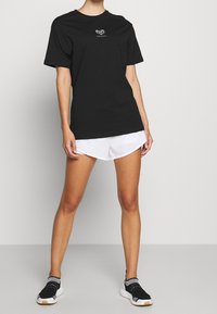 Černé bavlněné tričko s krátkým rukávem, s bílým geometrickým logem na hrudi. V kombinaci s bílými šortkami a černými sportovními botami.