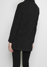 Manteau noir texturé avec un col à revers, manches longues et coupe droite. Présente une finition lisse et arrive à la hauteur des hanches.