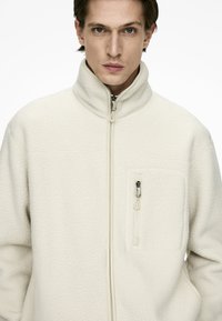 White fleece jacket with high collar, full zip, and chest pocket. Textured surface with a smooth zipper and matching pulls.