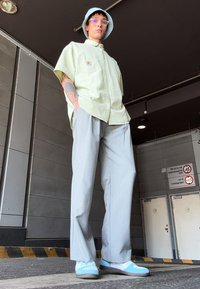Groen geruite kortemouwenshirt, grijze wijde broek, lichtblauwe sneakers en een lichtblauwe bucket hat, staand tegen een moderne achtergrond.