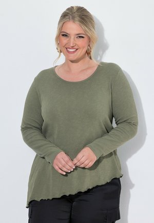 Femme souriante aux cheveux blonds porte un haut à manches longues vert olive avec un ourlet asymétrique et un pantalon noir, debout sur fond blanc.