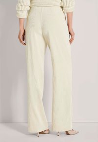 Persoon in crème-kleurige corduroy wijde broek, beige hoge hakken en een crèmekleurige gebreide trui, gezien van achteren.