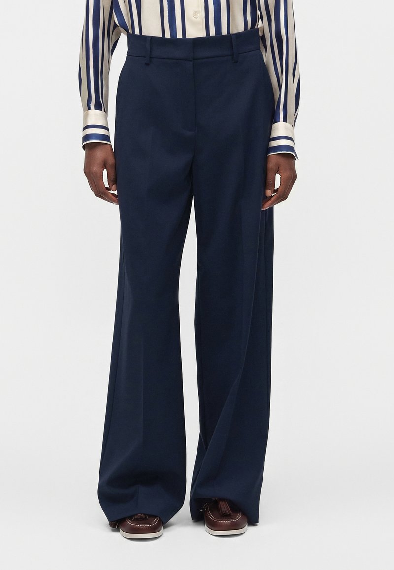Personne portant un pantalon large bleu marine assorti à une chemise boutonnée rayée verticale crème et bleu marine et des chaussures marron.