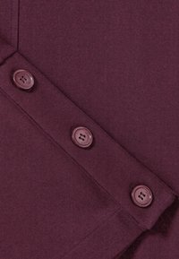 Tissu bordeaux avec une texture douce, présentant trois boutons ronds. Les boutons sont brillants avec deux trous, positionnés sur une couture diagonale.