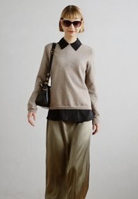 Suéter beige con un collar negro, superpuesto a una camisa negra, combinado con una falda de satén verde oliva. Los accesorios incluyen gafas de sol negras y un bolso.