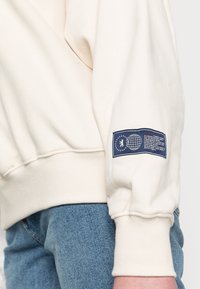 Persona con sudadera color beige claro con un parche azul en la manga y jeans de mezclilla azul, mostrada desde la mitad del torso hasta la muñeca.