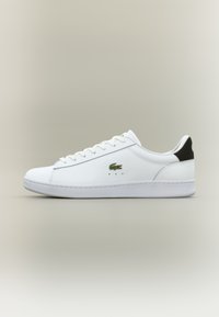 Bijele kožne tenisice s glatkom površinom, s zelenim izvezenim logotipom krokodila, ravnim bijelim potplatom i crnim naglaskom na peti.