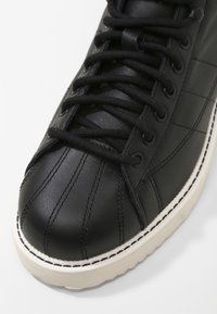 Black leather sneaker with a round toe, textured upper, prominent stitching details, and a white rubber sole. Features black laces and eyelets.