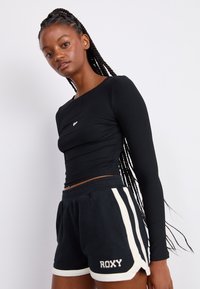 Camiseta de manga larga negra con textura acanalada que presenta un pequeño logo, combinada con pantalones cortos negros con rayas laterales blancas y detalles de logo, mostrando un diseño casual.