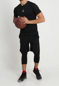 Svart träningsskjorta med en vit logotyp, kombinerad med svarta shorts och caprilånga leggings. Håller en brun basketboll och bär svarta skor.