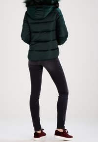 Giacca puffa verde con cuciture orizzontali e cappuccio foderato in pelliccia, abbinata a jeans skinny scuri e sneakers in suede bordeaux.