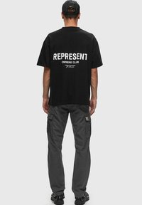 Camiseta negra con texto grande en blanco "REPRESENT" y "OWNERS' CLUB" en la parte de atrás, acompañada de pantalones cargo grises con bolsillos laterales.
