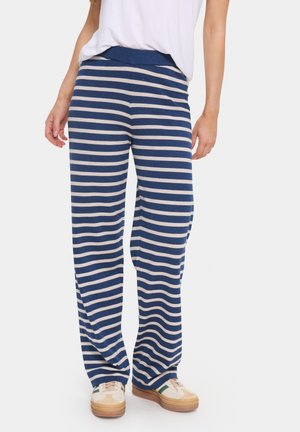 Onderlichaam van een persoon die blauw-witte horizontaal gestreepte wijde broek draagt met witte sneakers en een wit topje in de broek gestopt.