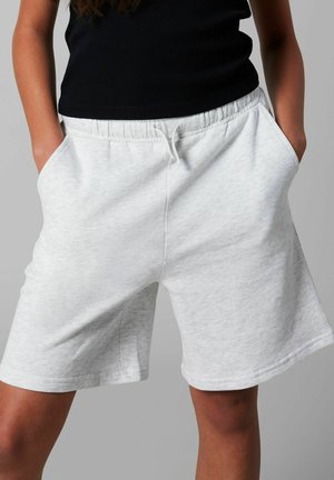 Femme portant un short gris clair à taille élastique et un haut noir sans manches, les mains dans les poches, sur un fond neutre.