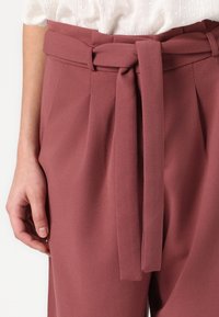 Pantalons ajustés rose avec une large ceinture, des plis à la taille et une texture lisse. Le tissu semble doux et léger.