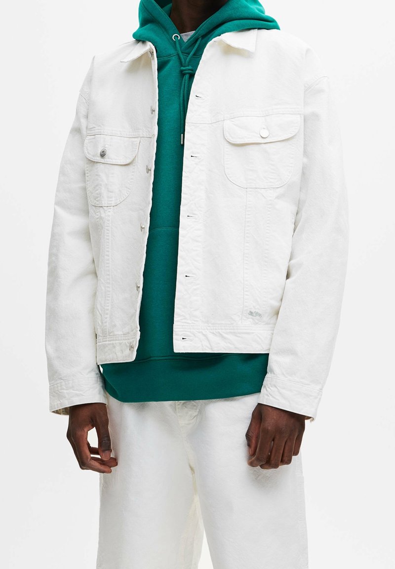 Vitt denimjacka med två fackfickor och silverdetaljer, över en grön hoodie. Passformen är lös och avslappnad, med en lätt textur.