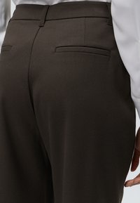 Gros plan d’un pantalon marron foncé sur mesure avec poches passepoilées à l’arrière, porté avec une chemise blanche, montrant une partie de la main sur le côté.