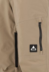 Veste beige avec une poche zippée sur le côté dotée d'une fermeture éclair noire, un tissu texturé et un logo en patch noir avec un design de montagne blanche.