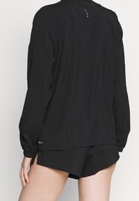 Top largo negro de manga larga con puños elásticos y dobladillo redondeado, que presenta un diseño sutil con pequeños detalles en la parte trasera. Combinado con pantalones cortos negros.