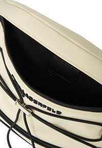 Innenraum einer beigen Nylon-Tasche mit schwarzer Fütterung, Reißverschlussverschluss und auffälligem Markenlogo. Enthält schwarze Akzentdetails und Beschläge.