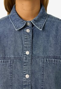 Blå denimskjorta med krage, silverfärgade tryckknappar, två framfickor och ett texturerat tyg med en lätt urblekt finish.