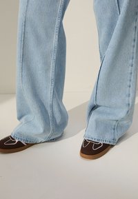 Jeans a gamba larga azzurro chiaro con orlo sfrangiato e scarpe marroni con cuciture rosa e design curvo sulla punta.