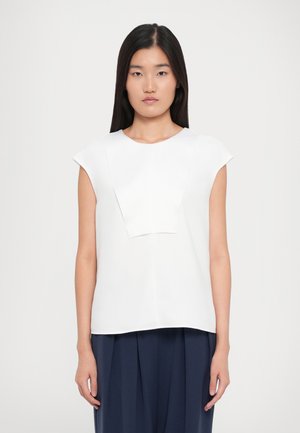 Giovane donna con capelli neri lisci indossa un top bianco senza maniche con un pannello frontale rettangolare e pantaloni a pieghe blu navy, in piedi contro uno sfondo bianco.