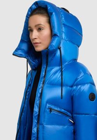 Modrá péřová bunda s velkou kapucí, vyrobená z lesklého materiálu. Má černý zip, kapsy a nášivku s logem na rukávu.