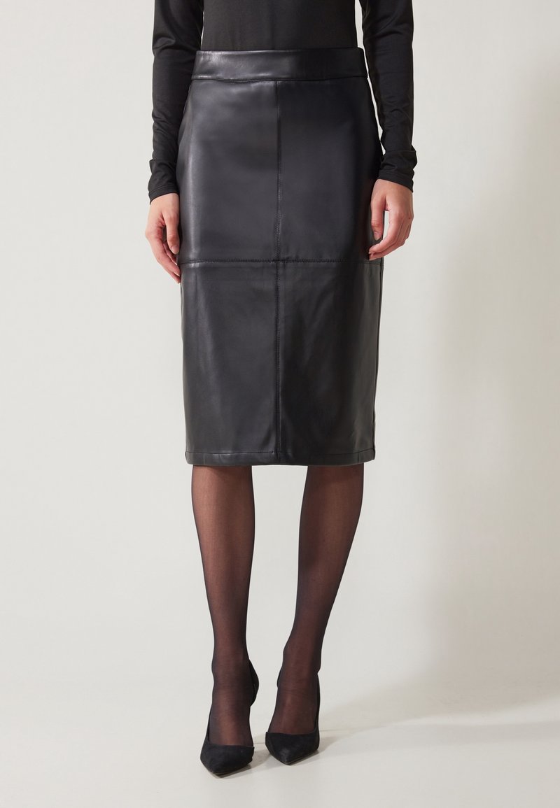 Kvinna i en svart läder knälång blyertskjol, genomskinliga svarta strumpbyxor, svarta högklackade skor och en långärmad svart topp mot en neutral bakgrund.