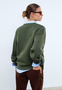 Sweatshirt vert olive avec un col rond, superposé sur une chemise à rayures bleu clair, associé à un pantalon marron en tissu texturé.