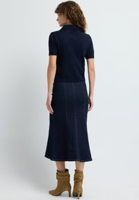 Kvinde med krøllet hår iført en marineblå kortærmet polo, en mørk denim midi-nederdel og afslappede beige ruskinds støvler med hæl, vendt væk.