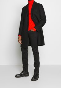 Uomo che indossa un cappotto nero, un dolcevita rosso, pantaloni neri e stivali di pelle neri, in piedi contro uno sfondo bianco semplice.