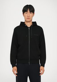 Svart zip-up hettegenser med snorhette, laget av glatt stoff. Har en diskret logo på brystet, ribbede mansjetter og nederkant.