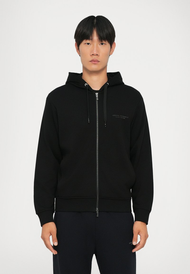 Svart zip-up hettegenser med snorhette, laget av glatt stoff. Har en diskret logo på brystet, ribbede mansjetter og nederkant.