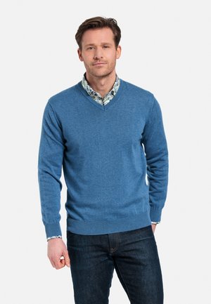 Blauwe v-hals trui van zachte stof, met ribbelmanchetten en -zoom, gedragen over een bloemetjesoverhemd. Donkere jeans complementeren de outfit.