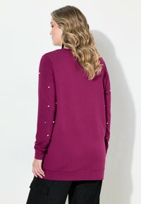 Dlouhý rukáv fialového topu s hladkou texturou, který má perlové ozdoby na rukávech a rovný lem.