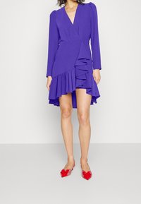 Lila långärmad klänning med inslag av omslagsdesign, med volangdetaljer och en hög-låg fåll, kombinerad med röda spetsiga skor.