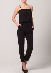 Combinaison noire avec fines bretelles, décolleté froncé, fermeture à boutons sur le devant, taille élastique et jambes fuselées avec poches. Texture de tissu lisse.