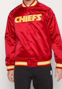 Giacca di raso rossa con logo "CHIEFS" giallo sul davanti. Presenta un colletto e polsini giallo e rosso a righe. Chiusura con bottoni a pressione.
