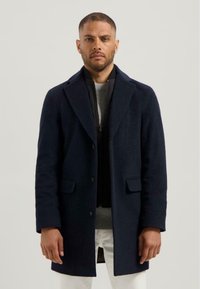 Marineblauwe wollen jas met notch-lapels, enkelvoudige sluiting en twee zijzakken, gelaagd over een gestructureerde grijze trui. Indrukwekkende silhouet.